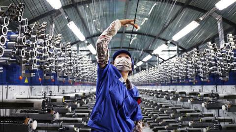 In a factory, a woman in a blue uniform and face mask reaches up with her right hand to hold a string above her head