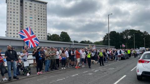 People with flags and banners stand protesting at the side of the road, watched by police