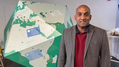 A man dressed in a dark jacket and red jumper stands in front of a large map of the world with labels pinpointing places ShelterBox has helped.