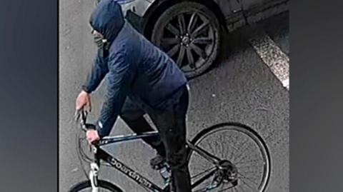 A man on a bike against a grey background. He is wearing a blue coat with the hood up and black trousers and is on a black bike with a white bar by the wheel.