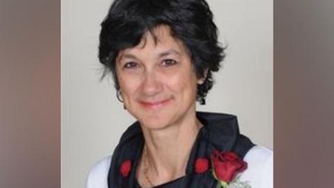 A grainy image showing Katie Thornburrow who has short dark brown hair and is wearing a white top with a black cowl collar, with a red rose pinned to her left shoulder. She is smiling.
