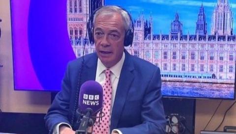 Reform UK leader, Nigel Farage sitting in a radio studio wearing a blue blazer, pink spotty tie, headphonea and talking into a microphone. 