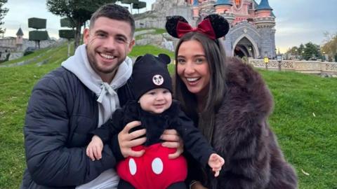 A man and woman wearing Minnie Mouse ears with a baby dressed as Mickey Mouse at Disneyland in front of the big pink castle