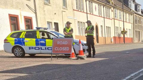 Two male police officers are standing by a police car in a street of terraced houses. They are all behind a ROAD CLOSED sign.