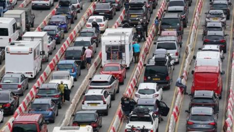 Cars and caravans lined up in rows of lanes. The cars are not moving. People are stood next to their stationary cars.