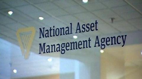 A glass door with a gold logo in the shape of an upside down triangle wit lines running through it. Blue lettering is printed on the glass saying National Asset Management Agency.