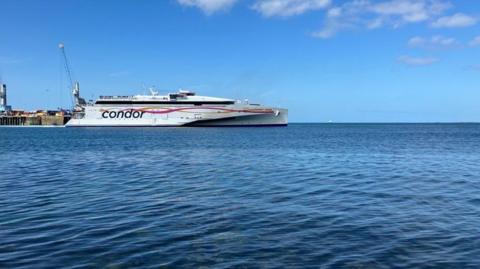 A white ferry with the Condor logo on the side in blue leaves a harbour on a sunny day.