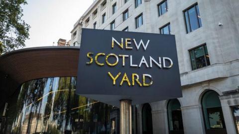 New Scotland Yard stands in front of a modern glass entrance and  stone building, which houses the headquarters of the Metropolitan Police.