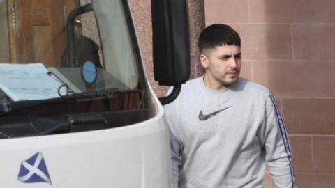 A man in his mid-20s with short black hair and stubble stands beside a white prison van. He is wearing a grey Nike sweatshirt.