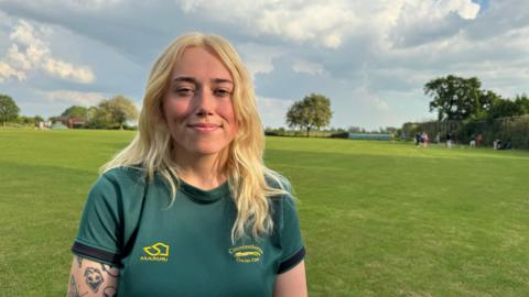 A woman with blonde hair wearing a green sports top standing in a cricket field with trees and blue skies behind her