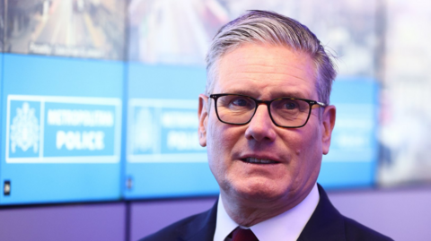 Close up of Prime Minister Keir Starmer standing in front of a screen displaying the Metropolitan Police logo (white writings on blue background)