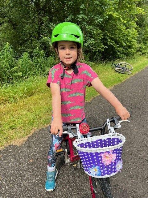 Young Eilidh Moyer in a bright green bike helmet, pink t shirt and blue trainers sits on her bike