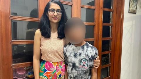 A woman wearing a colourful skirt and beige top stands in front of a door with her arm around her son. He is wearing a black and white t-shirt and his face has been blurred to protect his identify.