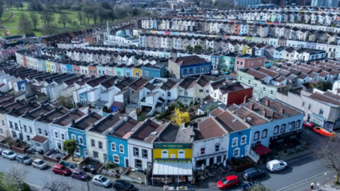 Birds eye view of a residential area in Bristol. There are multiple terraced houses and cars parked in front of the houses. All of the houses have been painted different colours - some blue, yellow, green, pink and white.