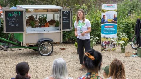 The back of people's heads as they watch a talk given by a member of staff at the Eden Project. They are standing outside and there is a stall behind them on wheels, serving soup