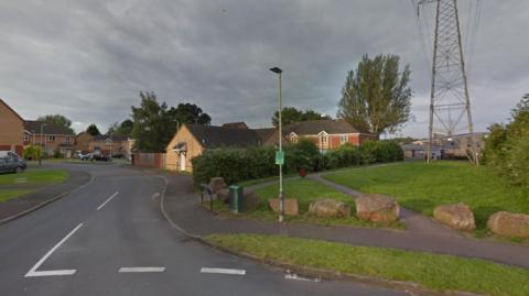 A residential road, Grenadier Road, on a sunny summer evening. Google Maps screenshot shows junction leading to road next to a green space with paths, big rocks and an electricity pylon.