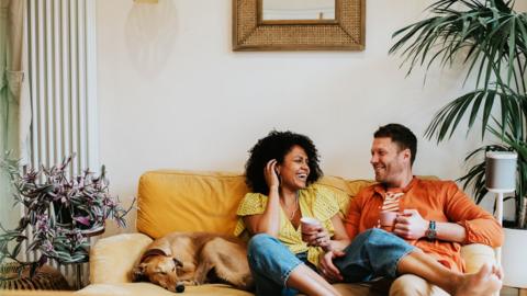 A man and a woman sit on a yellow sofa alongside their dog. They are laughing and smiling at each other whilst the dog sleeps.