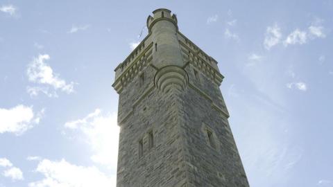 A view up towards the top of the MacDonald Monument. It is a tall, stone tower with narrow windows, and faux turrent and battlements. It is a sunny day.