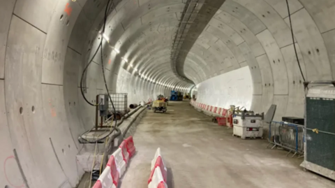 An older image showing the unfinished tunnel with grey walls, machinery and red and white barriers