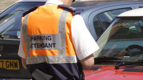 A parking attendant wearing a high visibility jackets and white shirt stands in front of two parked cars with his back to the camera