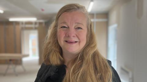 Jane Holmes is smiling at the camera in a building that is being built. She has long blonde hair.