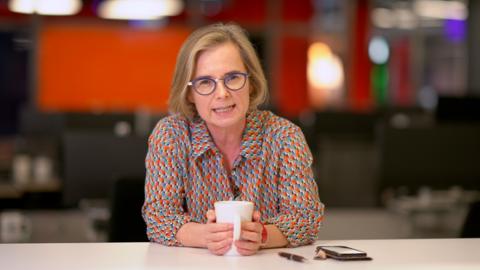 BBC Wales correspondent Felicity Evans, a woman with shoulder length light hair, sits at a desk wearing a brightly coloured shirt and holding a mug.