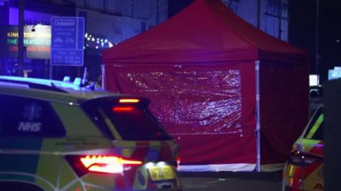 A red police tent set up in the road where the incident took place. Two emergency vehicles are parked in front of the tent. It is dark.