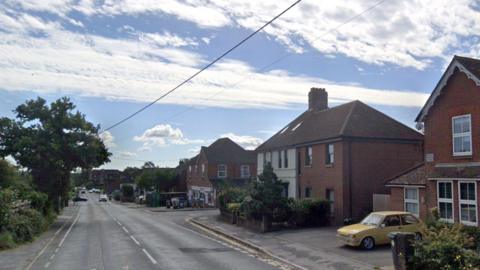 Winchester Road in Bishops Waltham. There are red brick houses lining the right of the road, with some greenery in the front gardens and there is an old fashioned yellow car parked in a driveway. Further down the road on the right is a village shop. To the left of the road are trees and more greenery and a traffic light can be seen in the distance.