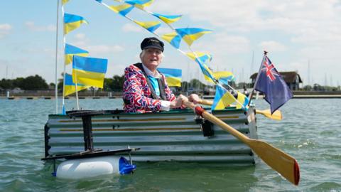 Major Mick, wearing a union jack suit and a black sea captain's peaked cap, sits in his tin bath boat, with a pair of oars, as he rows it on water. The boat is decorated with a union flag, a blue ensign and blue and yellow bunting.