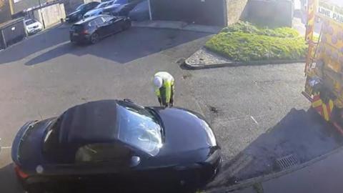 Black car sits in a large bay marked with white lines drawn around it. The car sits in a cul-de-sack with other vehicles. A contractor works on the ground. 