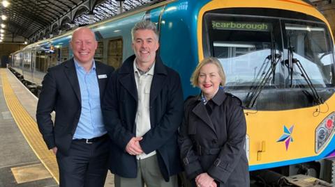 Two men and a woman stand in front of a train with the Transpennine Express logo on the front and Scarborough on the destination board.