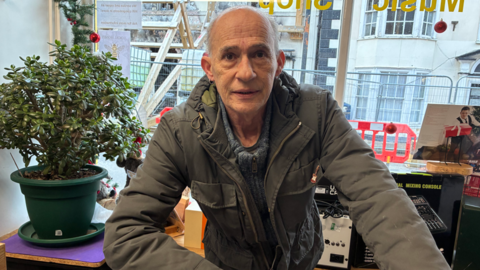 Man in a green jacket over a light blue jumper inside a building. He is leaning on a counter looking at the camera
