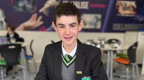 Boy with dark hair wearing black school blazer with green emblem, grey jumper, white shirt and green and black tie. 

He is sat at a grey circular desk in a classroom, a robot made from Lego and a laptop in front of him. 