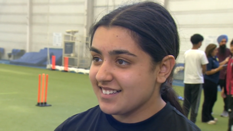 Millie can only be seen from the shoulders up, but she is smiling with her teeth and looking off to the side. She has long black hair which has been tied up into a low ponytail with a middle part. She is wearing a black crew neck sports T-shirt. Out of focus behind her are other players standing together and plastic orange cricket stumps on a green astro floor.
