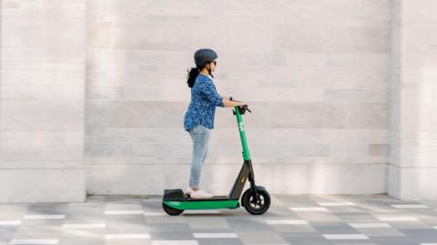 A woman with black hair and wearing a blue blouse and jeans rides on a green electric scooter with the Bolt logo on the frame.