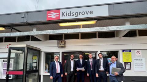 A group of six men wearing suits are stood on the station concourse of Kidsgrove Railway Station. There is a phone box to the left hand side of the image, and railway signage on the building behind them.
