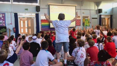 Crowds of children, some with white and red England shirts, have their backs turned to the camera, watching a projector. One child stands up with his arms wide gesticulating
