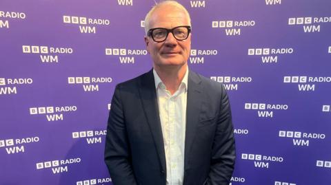 Mayor Richard Parker, a man with short white hair wearing glasses, a black suit jacket and a white shirt, stands in front of a purple war with BBC Radio WM branding on it.