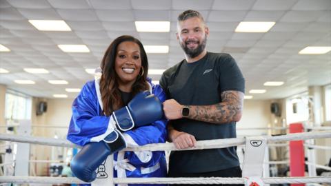 Alison Hammond and Tony Bellew stand on the edge of a boxing ring. Alison is wearing a blue boxing glove and fist pumping Tony.