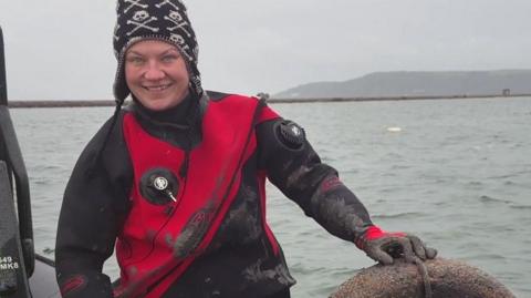 Mallory Haas sitting on the edge of a boat holding a tyre which has been retrieved from the seabed in Plymouth Sound. She is wearing a black and red wetsuit and a black woolly hat with a white skull pattern. She is smiling widely. The sea and the sky are both quite grey.