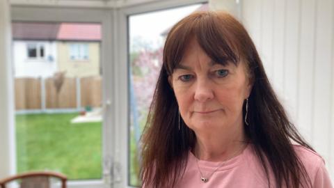 Chris Adamson, a woman with long brown hair, standing in a conservatory with a serious look on her face. She is wearing a pink top and necklace. A lawn, wooden fence and neighbouring houses can be seen through glass doors.