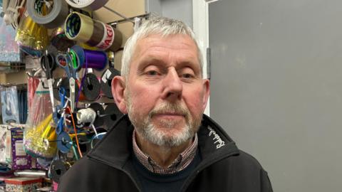 A head and shoulders image of Bill Bulstrode. He is standing in a shop wearing a black jacket over a jumper and a shirt. He is looking directly into the camera.