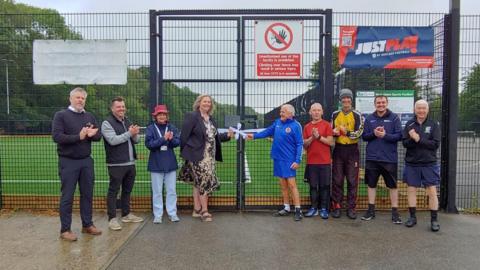 A group of people stood in front of a fence and gates at an astroturf football pitch. There are nine people, dressed mostly in football attire, but some in smart dress, and most of them are clapping.