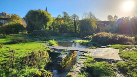 A low lying stream and basic bridge seen on a sunny day. Trees and a church spire can be seen in the background.