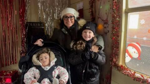 A woman in a black coat and white hat with a girl in a black coat and black hat and a girl in a wheelchair in a pink coat
