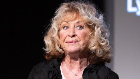La Plante pictured at an event in 2021, looking away from the camera with a pensive expression on her face, wearing a black top and big blonde curls.