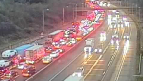 Both sides of the M25 at Brentwood in Essex. One side has a large queue of traffic, with most of the vehicles - including cars and lorries - glowing red from brake lights. On the other side, traffic is flowing normally.