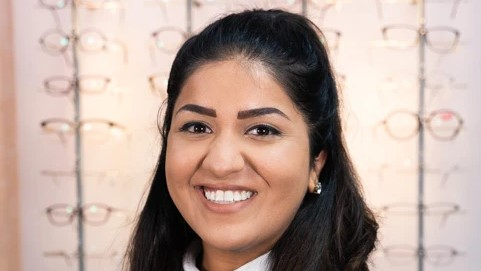 A portrait picture of Aashika Patel. She has long, black hair scraped back from her forehead. She is smiling at the camera. Behind her, there are four vertical racks with glasses displayed on them. 