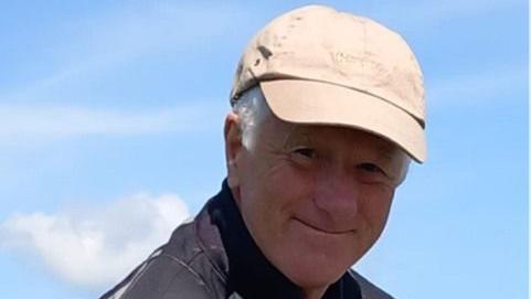 Richard is wearing a cream hat.
He is smiling at the camera with blue clouds behind him and one fluffy white cloud. 
He is wearing a black raincoat and the photo is cropped from the shoulders up.