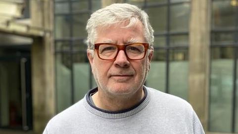 A man with light grey hair and short stubble looks at the camera with a neutral expression. He wears a light grey sweater and orangey-brown square glasses. There are glass panels on a building in the background.